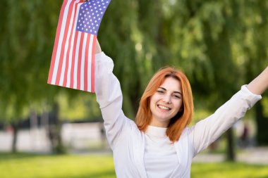 Mutlu kızıl saçlı kadın yazın açık havada ABD bayrağını tutuyor. Amerika 'nın bağımsızlık gününü kutlayan pozitif kız. Uluslararası Demokrasi Günü.