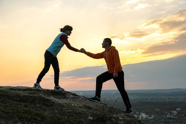 Man and woman hikers helping each other to climb stone at sunset in mountains. Couple climbing on high rock in evening nature. Tourism, traveling and healthy lifestyle concept.