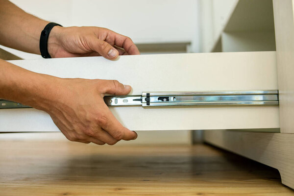 Close up of carpenter hands installing wooden drawer on sliding skids in contemporary cupboard cabinet.