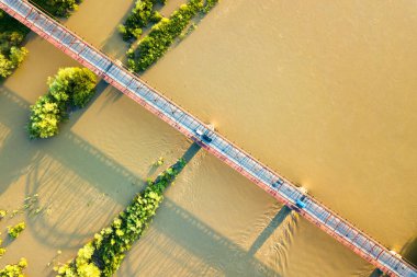 Yeşil kırsal bölgedeki çamurlu geniş nehir üzerinde uzanan dar bir yol köprüsünün havadan görünüşü.