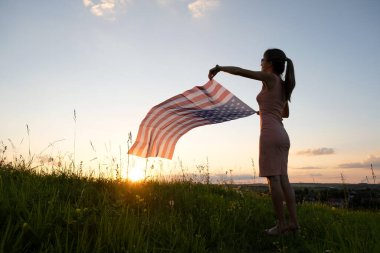 Gün batımında ABD bayrağıyla dışarıda poz veren mutlu genç bir kadının arka görüntüsü. Amerika 'nın bağımsızlık gününü kutlayan pozitif kız. Uluslararası Demokrasi Günü.