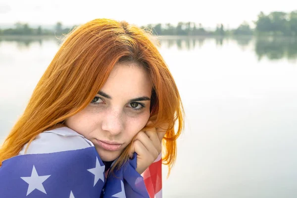 Portrait of sad red haired girl with USA national flag on her shoulders. Young woman celebrating United States independence day.