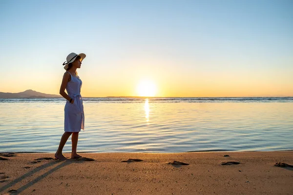 Hasır şapkalı ve elbisesi olan genç bir kadın gün batımında kumsalda tek başına yürüyor. Yalnız kız, sakin okyanus yüzeyinde tatil gezisinde ufka bakıyor..