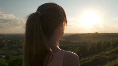 Genç, rahat bir kadın yeşil alanda duruyor ve akşam doğasında gün batımını seyrediyor. Rahatlama ve meditasyon kavramı.
