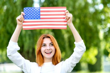 Mutlu kızıl saçlı kız kafasının üstünde Amerikan bayrağı sallayarak yaz parkında dikiliyor. Birleşik Devletler 'in bağımsızlık gününü kutlayan pozitif genç bir kadın. Uluslararası Demokrasi Günü.