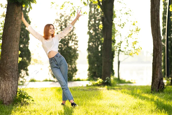 身穿白衬衫和蓝色牛仔裤的红头发年轻女人站在夏季公园里享受着温暖的天气 — 图库照片
