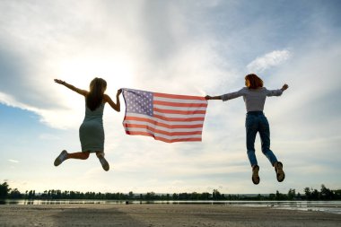 ABD bayrağı taşıyan iki genç arkadaş göl kıyısında birlikte zıplıyorlar. Vatansever kızlar Amerika 'nın bağımsızlık gününü kutluyor.