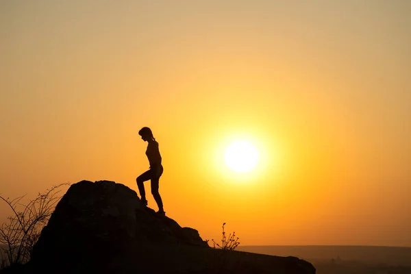 Silhouette of a woman hiker climbing up a big stone at sunset in mountains. Female tourist on high rock in evening nature. Tourism, traveling and healthy lifestyle concept.