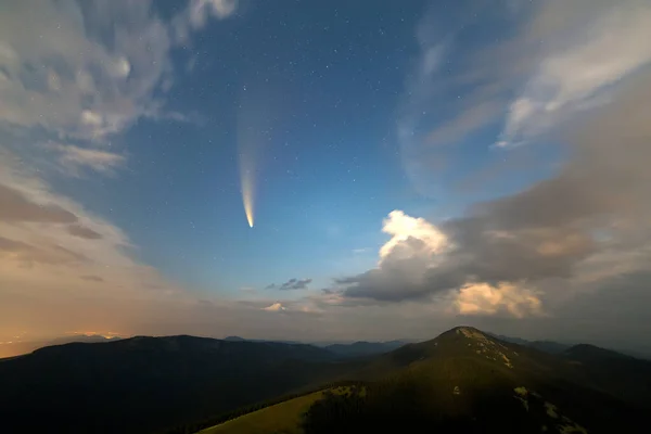 Summer night in mountains with starry dark blue cloudy sky and C/2020 F3 (NEOWISE) comet with light tail.