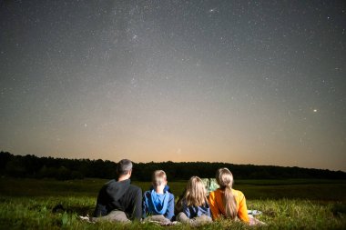 Gece tarlasında dinlenen mutlu bir aile bir sürü parlak yıldızlı karanlık gökyüzüne bakıyor. Ebeveynler ve çocuklar meteor yağmurunu izliyorlar.