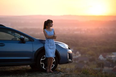 Mavi yazlık elbiseli mutlu kadın sürücü arabasının yanında sıcak bir akşamın tadını çıkarıyor. Seyahat ve tatil kavramı.