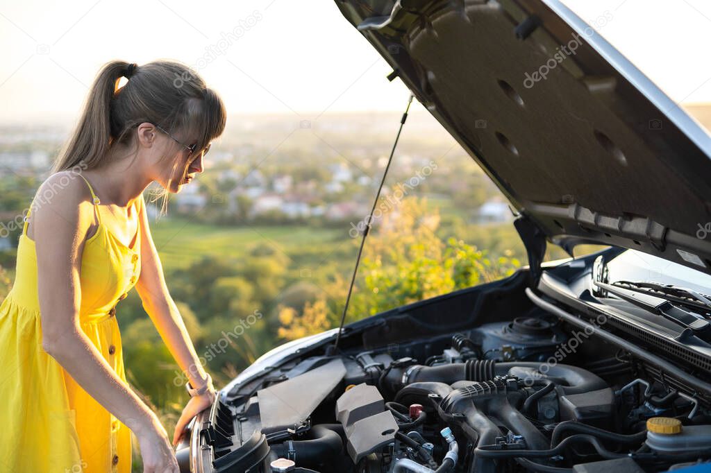 Puzzled female driver standing near her car with popped up hood looking at broken engine.