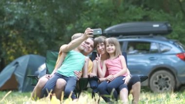 Aileler ve çocukları birlikte kamp alanında oturup cep telefonlarıyla selfie çekiyorlar..