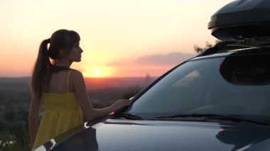 Yazlık elbiseli mutlu genç kız arabasının yanında duruyor ve günbatımının tadını çıkarıyor. Seyahat ve tatil kavramı.