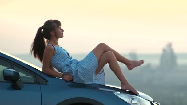暖かい夏の日を楽しむ彼女の車のフードに横たわって青いドレスの幸せな若い女性ドライバー 旅行と休暇のコンセプト — ストック動画