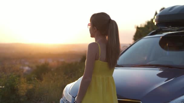 彼女の車の近くに立っている夏のドレスで若い女性のドライバーは 自然の夕日の景色を楽しんで 旅行と休暇のコンセプト — ストック動画