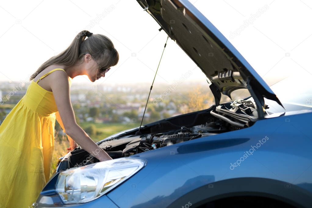 Puzzled woman driver standing near her car with popped up hood inspecting broken engine.