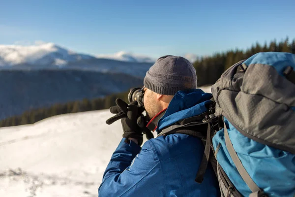 冬の寒い日に青空の下で雪の谷や木製の山の峰の風景の写真を撮るバックパックとカメラと暖かい服でハイカーの男の観光客の写真家 — ストック写真