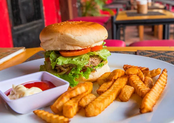 Big burger with potato on white plate. Juicy beef burger with salad and tomato with ketchup. Rich meal for children. Eating hamburger in cafe or restaurant. Simple snack served for morning breakfast