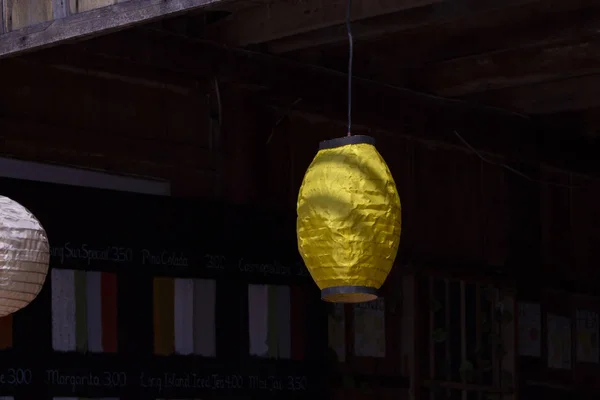 Yellow paper lantern in Chinese style, outdoor decor. Yellow paper lamp on dark background. Rustic handmade decoration on countryside house. Summer home exterior idea. Soft paper lantern in sunshine