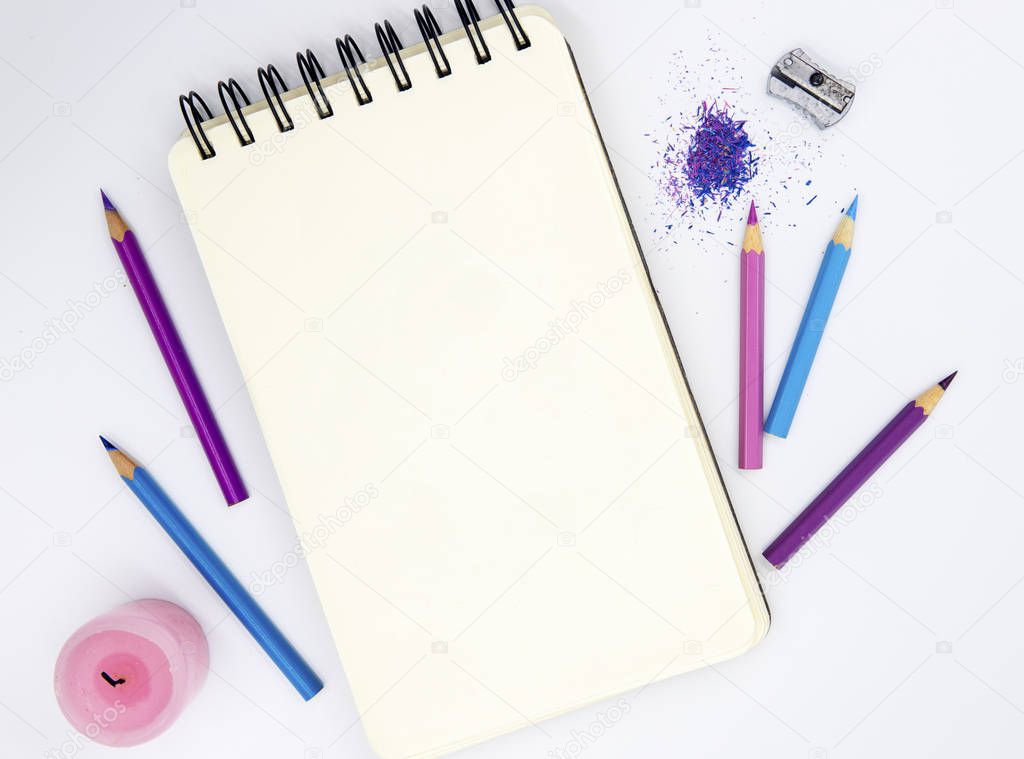 Empty sketchbook, pink violet pencils, pink candle on white background. Artistic table top view photo. Creative hobby workplace. Blank sketchpad and crayons flat lay. St Valentine logo drawing mockup