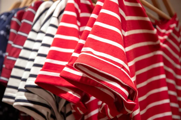 Striped shirts on hangs for sale in shop. Red and white polo on wooden hanger. Summer seasonal wear in department store. Unisex apparel for warm weather. Sale in shopping mall. Nautical style t-shirts