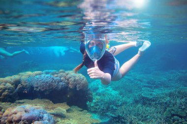 Denizaltında şnorkelle yüzen genç bir kadın. Şnorkel baş parmağını suyun altında gösterdi. Yüzü tamamen şnorkelli bir kadın. Mercan resiflerinin sualtı fotoğrafı. Tropikal adada yaz tatili. Deniz kenarı etkinliği