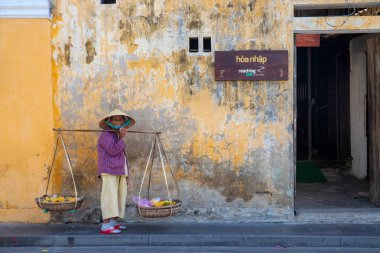 Hoi An, Vietnam - 28 Temmuz 2019: Vietnam şapkalı yaşlı kadın sırık taşıyor, arka planda tarihi bir duvar var. Vietnamlı konik şapkalı gülümseyen kadın.