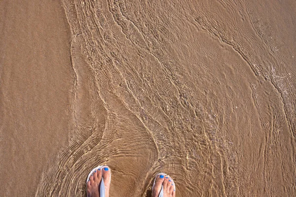 Παραλία άμμο και γυναικεία πόδια σε θάλασσα κύμα κορυφή άποψη. Χαλαρωτική φωτογραφία της αμμώδους παραλίας και καθαρό θαλασσινό νερό. Γυναικεία πόδια σε σανδάλια — Φωτογραφία Αρχείου
