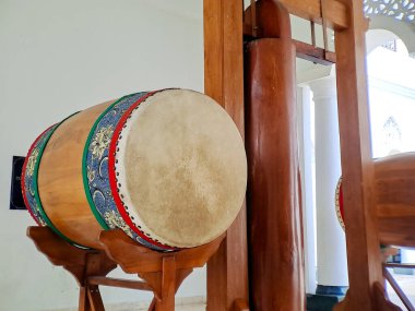 A large, traditional drum rests on a wooden stand inside a building with white walls.  