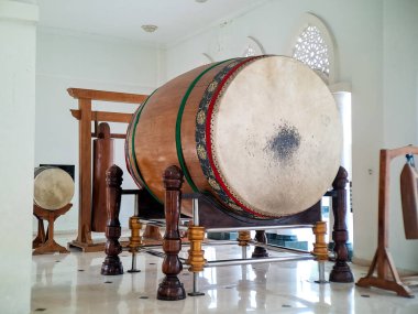 An antique wooden drum rests on its ornate stand inside a bright, white-walled building interior.