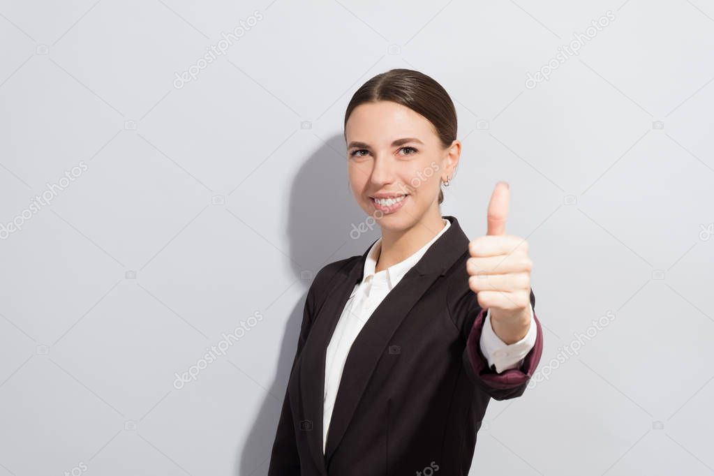 Young good looking business woman showing thumb up against grey background. Fashion style photoshoot with hard light source