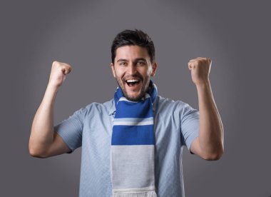 Yumruklarını gri arka plan üzerinde izole başını tutan eşarp mavi ile genç sakallı spor fan portresi. Takımınızı desteklemek