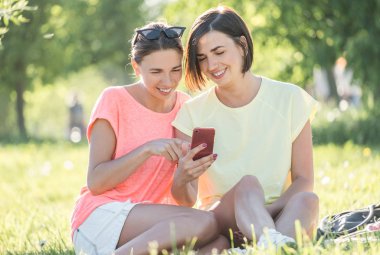 Açık portresi iki neşeli kız çimenlerin üzerine oturmuş, smartphone kullanarak ve iyi zaman birlikte parkta güneşli gün olması..