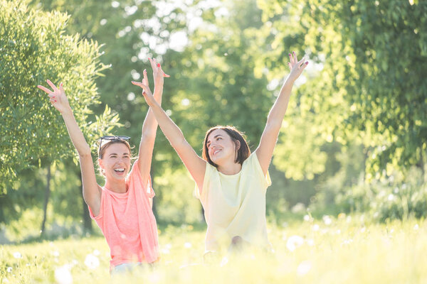 Outdoor portrait of two cheerful girls sitting on lawn with hands up in the air in park at sunny day. Young women spending good time together