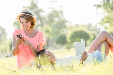 Çimenlerin üzerine oturmuş, kitap okumak, müzik Smartphone cep telefonu ile yeşil park güneşli sıcak gününde iki neşeli kız. Açık zaman yaz gün harcama