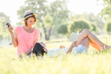 Çimenlerin üzerine oturmuş, kitap okumak, müzik Smartphone cep telefonu ile yeşil park güneşli sıcak gününde iki neşeli kız. Açık zaman yaz gün harcama