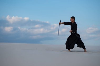 Geleneksel Japon giysili odaklı adam dövüş sanatları bir Japon kılıç - günbatımı arka plan mavi gökyüzü ve bulutlar üzerinde sırasında bir katana ile çölde pratik.