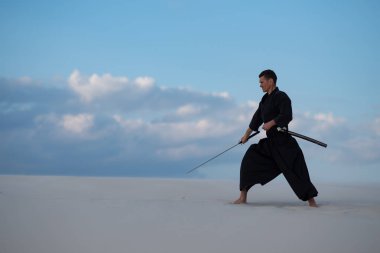 Geleneksel Japon giysili odaklı adam dövüş sanatları bir Japon kılıç - günbatımı arka plan mavi gökyüzü ve bulutlar üzerinde sırasında bir katana ile çölde pratik.