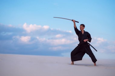 Geleneksel Japon giysili odaklı adam dövüş sanatları bir Japon kılıç - günbatımı arka plan mavi gökyüzü ve bulutlar üzerinde sırasında bir katana ile çölde pratik.