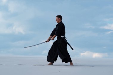 Mutlu adam, geleneksel Japon elbiseleri, dövüş sanatları bir Japon kılıç - günbatımı arka plan mavi gökyüzü ve bulutlar üzerinde sırasında bir katana ile çölde pratik.