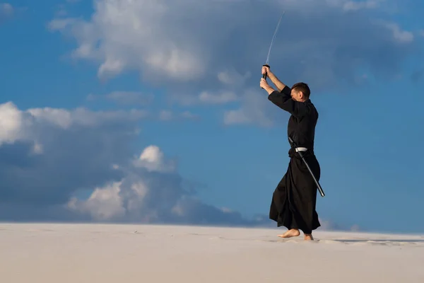 Resolute man, in traditional Japanese clothes, with raised sword, katana, is training martial arts in desert during sunset - samurai on the blue cloudy sky background.