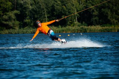 Wakeboarder, turuncu gömlek ve kask, sıçraması ise güneşli bir günde bir kablo parkta eğitim bulutu rides. Açık içinde aktif bir tatil.