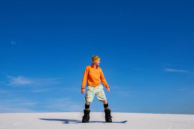 Güneşli bir gün, kum yatılı çölde. Adam mavi gökyüzünün karşı bir snowboard üzerinde kum tepeleri üzerinde süzülüyor. Geniş açı, yan görünüm.