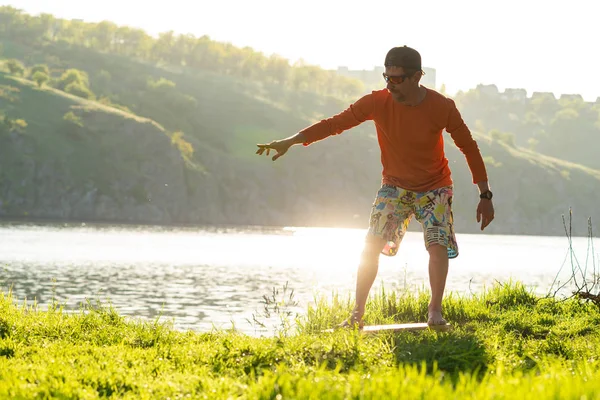 Bearded man is training on the balance board on a green meadow next to river in the rays of summer sun. Back light, front view.