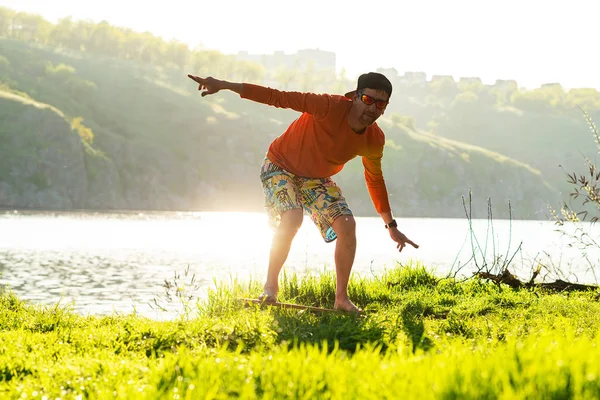 Bearded man is training on the balance board and having fun on a green meadow next to river in the rays of summer sun. Back light, front view.