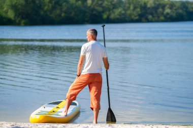 Yetişkin adam güneşli sabahı Sup kurulu plaj yanındaki bir kürek ile ayakta duruyor. Yukarı raket yatılı - stand harika Aktif istirahat tatil sırasında. Arkadan görünüm.