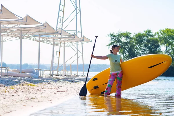 Mutlu kadın sahilde elinde Sup kurulu ile ayakta ve mesafe içine arıyorum. Yukarı raket yatılı - stand tatil sırasında harika Aktif istirahat.