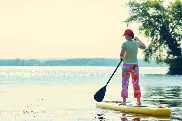 Yetişkin kadın bir Sup tahta üzerinde güneşli sabahı ölü olarak yatıyor. Yukarı raket yatılı - stand harika Aktif istirahat tatil sırasında. Arkadan görünüm.