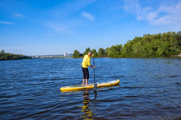 Adult man is training to sail on a SUP board on sunny morning on cityscape background. Stand up paddle boarding - awesome active recreation during vacation. Wide angle.